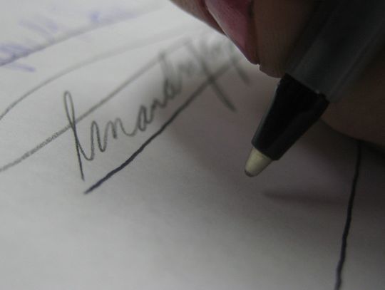 close image of a ballpoint pen being held over a piece of paper by someone about to sign their name. Other signatures are visible above where the pen is being held.