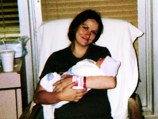 A photo of a woman with dark hair wearing dark clothes holding a baby wrapped in a white blanket and wearing a knit cap. The woman is looking at the camera.