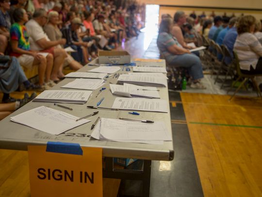photo of a gymnasium where a public meeting is being held. In the foreground, a rectangular table with several stacks of papers and pens scattered upon it with a sign saying "SIGN IN" in all capital large black letters on orange paper is taped to the table. Several rows of people in the background