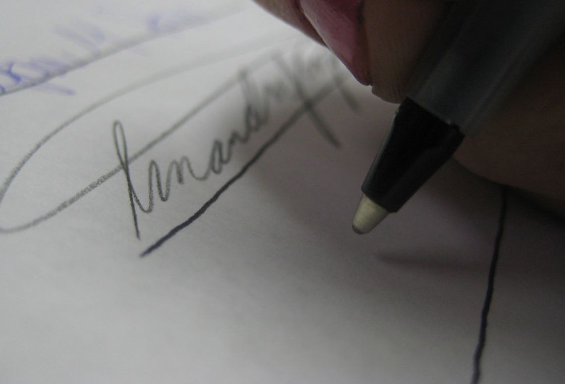 close image of a ballpoint pen being held over a piece of paper by someone about to sign their name. Other signatures are visible above where the pen is being held.