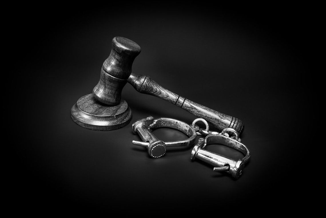 Black and white stock image of a judge's gavel and a pair of old-fashioned handcuffs set against a black background