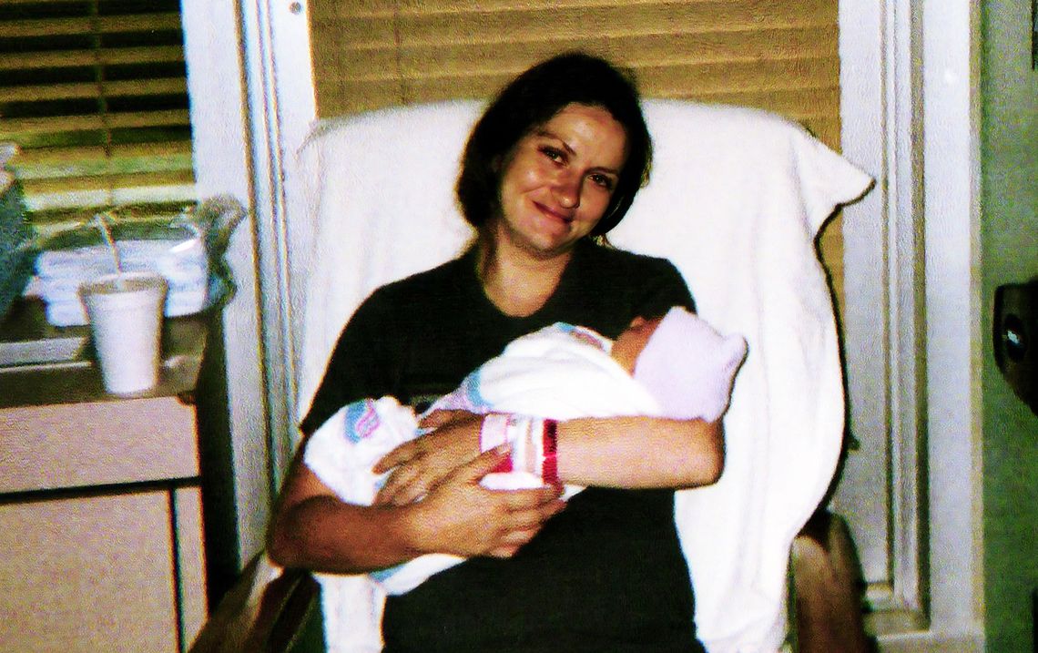 A photo of a woman with dark hair wearing dark clothes holding a baby wrapped in a white blanket and wearing a knit cap. The woman is looking at the camera.